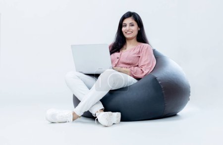 Photo for Indian female with laptop sitting on Bean bag on isolated white background and smiling looking towards the camera - Royalty Free Image