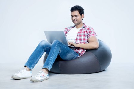 Photo for Indian man working on laptop while sitting on bean bag on isolated background - Royalty Free Image