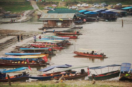 Téléchargez les photos : KANCHANABURI, THAÏLANDE - 16 AVRIL 2016 : Un groupe non identifié de bateaux à longue queue emmène les touristes le long de la rivière Songkhalia près du pont Mon ou du pont Uttamanusorn. Situé Amphoe Sangkhlaburi, Kanchanaburi Province, Thaïlande. - en image libre de droit