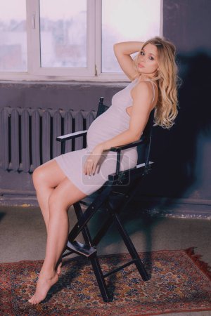 Téléchargez les photos : Une image saisissante d'une femme enceinte dans une robe gris clair moulante, assise élégamment dans une chaise de réalisatrice noire près d'une fenêtre. Réflexion dans le miroir. - en image libre de droit