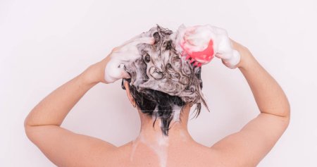 Señora con espuma en el pelo cuida su cuero cabelludo con masajeador rosa sobre fondo blanco.
