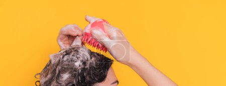 Dame mit Schaum auf dem Kopf pflegt ihre Kopfhaut mit rosa Massagegerät vor gelbem Hintergrund.