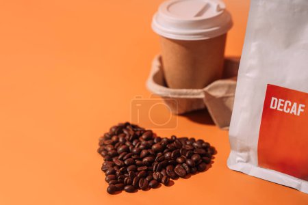 Foto de Granos de café en forma de corazón sobre un fondo anaranjado junto al envase con texto descafeinado y una taza de papel en una bandeja. - Imagen libre de derechos