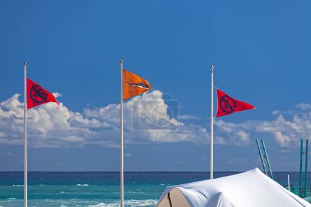 Foto de Tres banderas de advertencia ondeando en el aire en el Plage des Roches Noires en Saint-Gilles les Bains durante el solsticio de verano. Las dos banderas rojas indican "no nadar" y "no surfear", mientras que la bandera naranja advierte sobre la posibilidad de tiburones en el wat - Imagen libre de derechos