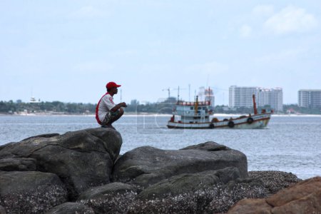 Foto de Hua Hin, Tailandia - 12 de agosto de 2007: Hombre tailandés en cuclillas sobre una roca mientras mira al horizonte. Detrás de él, un barco pesquero se va al mar.. - Imagen libre de derechos