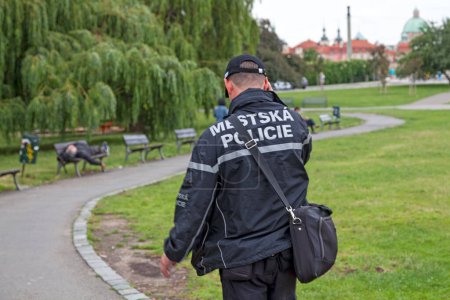 Foto de Praga, República Checa - 14 de junio de 2018: Oficial de policía de la Mestska Policie caminando por un parque mientras habla por su teléfono móvil. - Imagen libre de derechos