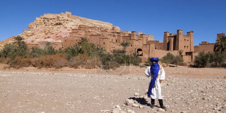 Foto de Ait Benhaddou, Marruecos - 30 de enero de 2019: Bereber con ropa tradicional frente a Ait Benhaddou, un pueblo fortificado a lo largo de la antigua ruta de caravanas entre el Sahara y Marrakech en el actual Marruecos. - Imagen libre de derechos