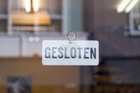 White an red sign hanging at the glass door of a shop saying in Dutch: "Gesloten", meaning in English "Closed".