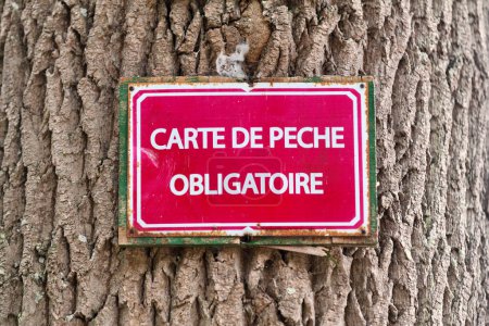 Foto de Cartel clavado en un tronco de árbol que dice en francés "Carte de peche obligatoire", que significa en inglés "Obliatory fishing card". - Imagen libre de derechos