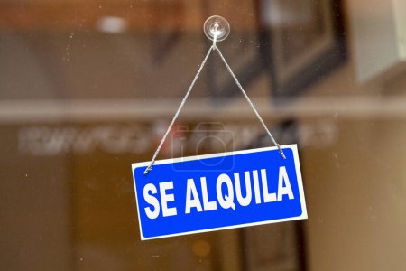 Blue sign hanging at the glass door of a shop saying in Spanish "Se alquila" meaning in English "To rent".