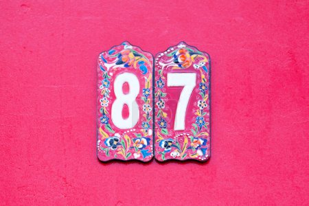 Colorful earthenware plate with a number 87 in its middle. The plate is stuck on the front of a house painted in strawberry.