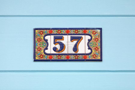Colorful earthenware plate with a number 57 in its middle. The plate is stuck on the front of a house painted in sky blue.