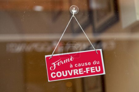 Red sign hanging at the glass door of a shop with written in French "Ferme a cause du couvre-feu", meaning in English "Closed due to curfew" in the middle.