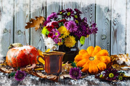 A bouquet of autumn flowers in a retro pot, orange pumpkins, autumn leaves, a vintage cup. Late autumn, November, the first snow. Still life on a snowy wooden background.