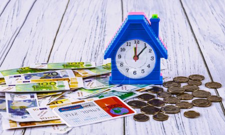 Foto de Home loan concept. Toy euro banknotes and cents, credit card, alarm clock in the form of a house on a blue background. Investments in construction - Imagen libre de derechos
