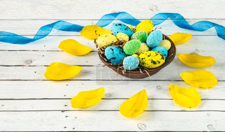 Plate with blue and yellow Easter eggs and tulip petals, blue ribbon on a light wooden background. Easter composition in blue and yellow colors. View from above