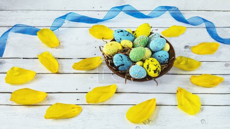 Téléchargez les photos : Assiette avec œufs de Pâques bleus et jaunes et pétales de tulipe, ruban bleu sur fond bois clair. Composition de Pâques en couleurs bleu et jaune. Vue d'en haut - en image libre de droit