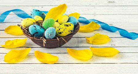 Téléchargez les photos : Assiette avec œufs de Pâques bleus et jaunes et pétales de tulipe, ruban bleu sur fond bois clair. Composition de Pâques en couleurs bleu et jaune. Vue latérale. - en image libre de droit