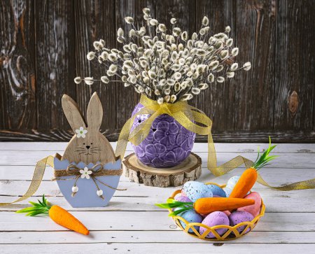 Foto de Conejo de Pascua con huevos de Pascua y zanahorias. Un ramo de ramitas de sauce en un jarrón como decoración. Composición festiva sobre fondo de madera. - Imagen libre de derechos