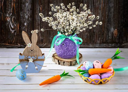 Photo for Easter bunny with Easter eggs and carrots. A bouquet of willow twigs in a vase as a decoration. Festive composition on a wooden background. - Royalty Free Image