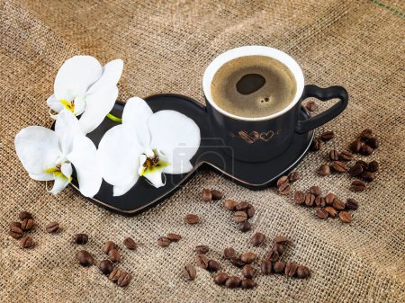 Photo for Black cup of coffee and white orchid flowers on a dark background. Coffee beans and burlap as decor. Romantic coffee composition.View from above - Royalty Free Image