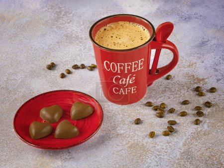 Foto de Una taza de café rojo con una cuchara, granos de café y chocolates en un plato rojo sobre un fondo claro. - Imagen libre de derechos