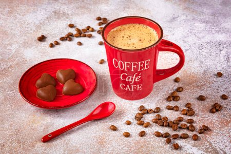 Foto de Una taza de café rojo con una cuchara, granos de café y chocolates en un plato rojo sobre un fondo claro. - Imagen libre de derechos