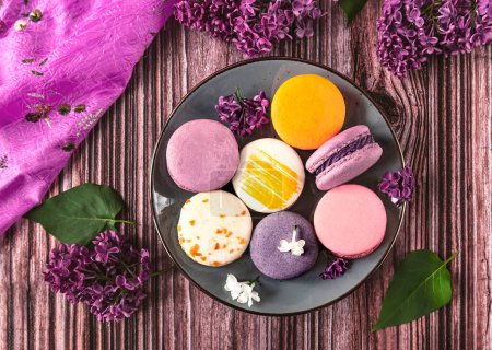 Photo for Plate with colorful French dessert macaroons on wooden background, top view. Purple lilac flowers as decoration - Royalty Free Image