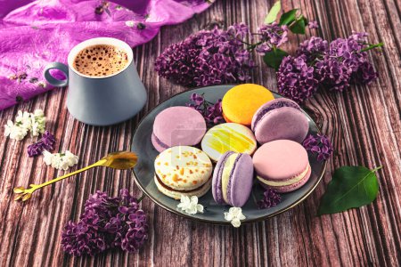 Photo for Colorful dessert macaroons on a plate with a spoon, gray cup of coffee on the table. Purple lilac as a decoration - Royalty Free Image