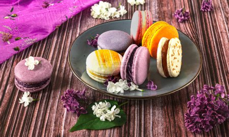 Photo for Plate with colorful French dessert macaroons on wooden background. Purple lilac flowers as decoration, side view - Royalty Free Image
