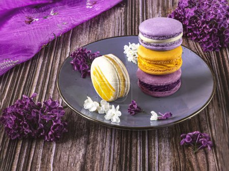 Photo for Composition with purple and yellow macaroons desserts on a plate and table. Lilac flowers as decoration. - Royalty Free Image