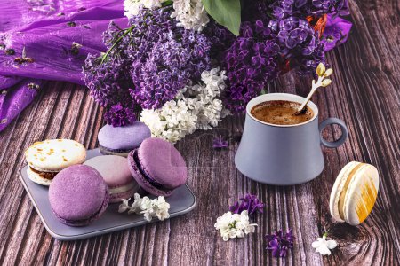 Foto de Un plato con un colorido postre francés de macarrones sobre un fondo de madera y una taza de café gris. Flores lila púrpura como decoración - Imagen libre de derechos