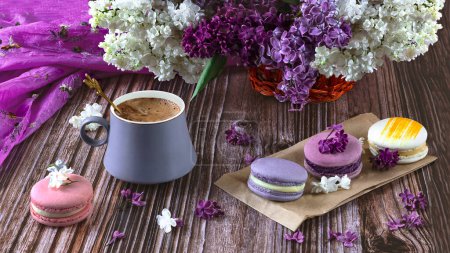 Photo for A row of colorful macaroon desserts on the table and a gray cup of coffee, side view. Purple bouquet of lilac flowers. - Royalty Free Image