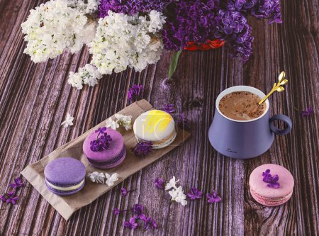 Photo for A row of colorful macaroon desserts on the table and a gray cup of coffee. Purple bouquet of lilac flowers. - Royalty Free Image
