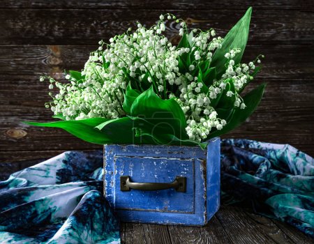 Téléchargez les photos : Un bouquet de lys des fleurs de la vallée dans une boîte vintage en bois bleu et un châle bleu sur un fond en bois. Vue d'en haut. - en image libre de droit