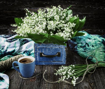 Foto de Un ramo de fragantes lirios del valle florece en una caja vintage azul, una taza de café azul y una bufanda sobre un fondo de madera. Primavera naturaleza muerta - Imagen libre de derechos