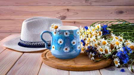 Foto de Una taza de café azul con margaritas, un ramo de margaritas y acianos, un sombrero de sol. Naturaleza muerta de verano con flores silvestres. - Imagen libre de derechos
