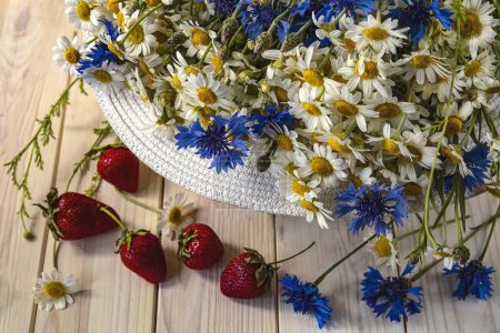 Photo for A bouquet of daisies and cornflowers in a sun hat. Summer still life with wildflowers and strawberries, top view - Royalty Free Image