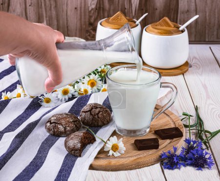 Foto de Un vaso de leche, una botella de leche y galletas con chispas de chocolate sobre un mantel sobre una mesa. Flores silvestres de verano como decoración, vista lateral - Imagen libre de derechos