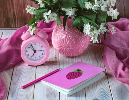 Foto de Bodegón de verano con un despertador rosa, un ramo de flores de jazmín, un cuaderno sobre la mesa. Hola concepto de verano - Imagen libre de derechos