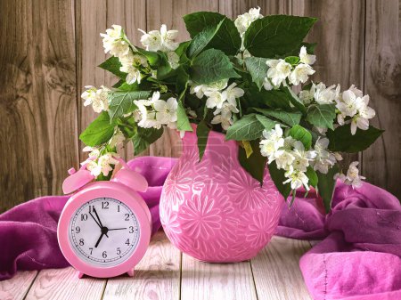 Foto de Reloj despertador rosa vintage y ramo de jazmín en jarrón rosa sobre fondo de madera. Concepto de verano mañana - Imagen libre de derechos