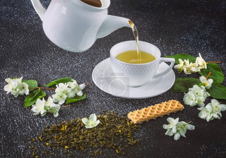 Foto de Té verde con jazmín en una taza blanca y tetera sobre un fondo oscuro. Flores frescas de jazmín y postre de gofre como decoración - Imagen libre de derechos