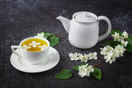 Foto de Té verde con jazmín en una taza blanca y tetera sobre un fondo oscuro. Flores frescas de jazmín como decoración - Imagen libre de derechos