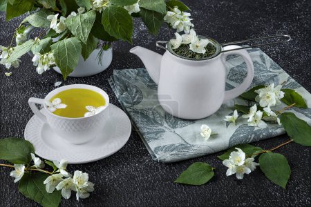 Foto de Naturaleza muerta con té verde en una taza blanca, tetera blanca sobre un fondo oscuro. Flores frescas de jazmín en un jarrón como decoración - Imagen libre de derechos
