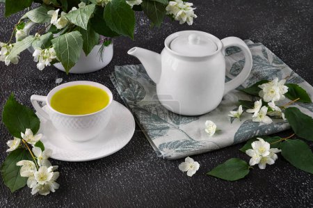 Foto de Té verde con jazmín en una taza blanca y tetera sobre un fondo oscuro. Flores frescas de jazmín en un jarrón como decoración - Imagen libre de derechos