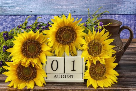 1. August auf einem weißen Holzkalender und schönen gelben Sonnenblumenblüten herum, ein Krug auf blauem Holzgrund.