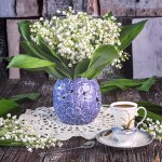 A bouquet of fresh lily of the valley flowers in a blue vase and a white cup of coffee on a dark wooden background. Spring still life with fragrant white flowers