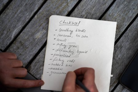 Téléchargez les photos : Gril writing a camping list in a piece of paper. writing a check list with a pencil and a phone - en image libre de droit
