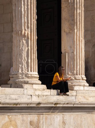 Téléchargez les photos : Nîmes, France - 4 octobre 2023 : jeune femme assise entre les piliers du temple romain Maison Carree dans la ville de Nîmes dans le sud de la France - en image libre de droit