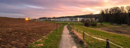 A beautiful golden hour panoramic scene in Switzerland: a walking path between fields near Katzensee lake and modern apartment buildings in Affoltern, Canton of Zurich